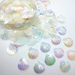 May include: A collection of translucent, iridescent seashell-shaped charms with a ribbed texture. Each piece features a small hole at the top for attachment. The charms display a pearlescent finish that shifts between soft pastel hues of pink, blue, and green.