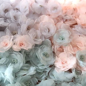 以下が含まれることがあります： 白、ピンク、淡い青色の繊細な布製花のコレクション。 各花には複数の花びらと小さなビーズの中心があります。 花は密接に配置され、テクスチャとレイヤー効果を作成します。