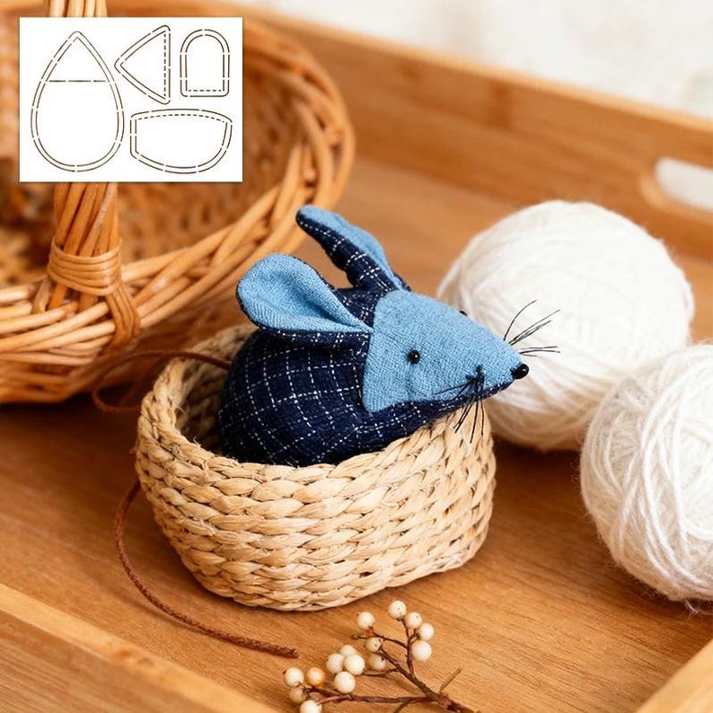 May include: A handcrafted fabric mouse with blue ears and a navy and blue plaid body sits in a small woven basket. Two white yarn balls and a paper pattern are in the background. The mouse has black whiskers and a brown tail.