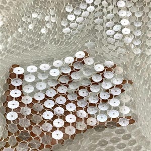 Può includere: Un tessuto bianco trasparente caratterizzato da una fitta griglia di paillettes argentate circolari e riflettenti. Il materiale è leggero e semitrasparente, con dischi metallici disposti in un motivo ripetitivo che crea una superficie.