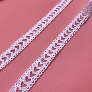 Könnte beinhalten: Zwei Streifen weißer Spitze mit einem herzförmigen Muster. Die Spitze ist filigran und detailliert, mit gleichmäßig verteilten Herzen an den Rändern. Der Hintergrund ist einfarbig rosa.