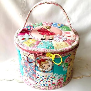 May include: A round, quilted fabric storage case with a handle, featuring a colorful patchwork design of floral and patterned textiles. The lid displays a central illustration of a girl in a red dress, while the side features a yellow and pink zipper.