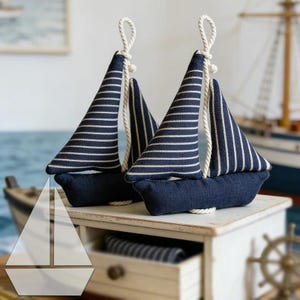Puede incluir: Dos adornos decorativos de veleros con cascos azul marino oscuro y velas de tela con finas rayas horizontales blancas. Cada velero tiene un lazo de cuerda blanca en la parte superior del mástil y está hecho de materiales de tela texturizada.