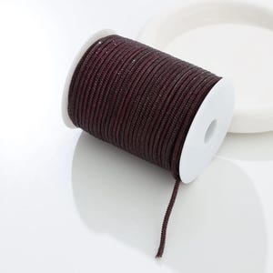 May include: A spool of dark burgundy cord with a subtle shimmer. The cord is tightly wound around a white plastic spool. A length of the cord extends from the spool, suggesting its use in crafts or jewelry making.