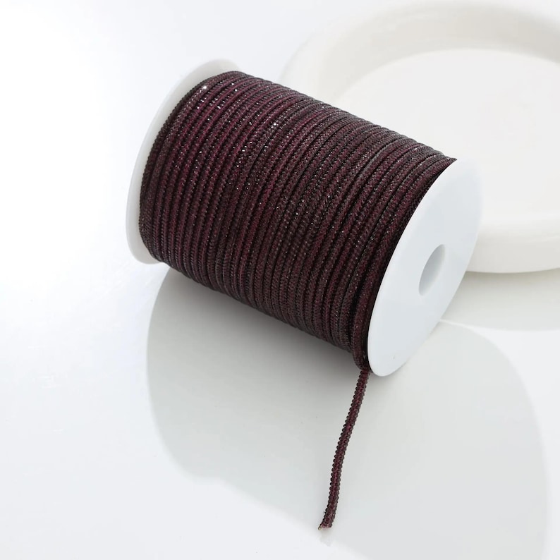 May include: A spool of dark burgundy cord with a subtle shimmer. The cord is tightly wound around a white plastic spool. A length of the cord extends from the spool, suggesting its use in crafts or jewelry making.
