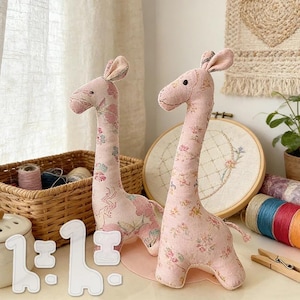 Può includere: Due giraffe di stoffa rosa con motivi floreali. Le giraffe hanno colli lunghi e piccole orecchie. Un cestino con rocchetti di filo e un modello di cucito sono in primo piano. Lo sfondo include un telaio da ricamo floreale.