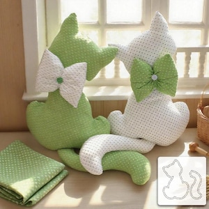 Puede incluir: Cojines hechos a mano con forma de gato en tela verde y blanca con lunares, cada uno con un lazo grande. Un cojín es verde con un lazo blanco, y el otro es blanco con un lazo verde. También se ve un cuadrado de tela verde doblado y un patrón de gato.