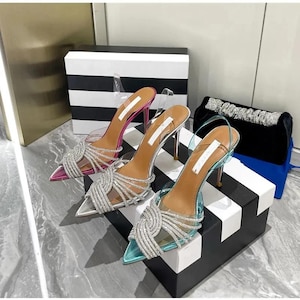 May include: Three pairs of high-heeled shoes in pink, silver, and teal. Each shoe has a pointed toe, clear straps, and a silver rhinestone design. The heels are thin and metallic. A black clutch with a silver beaded strap is also visible.