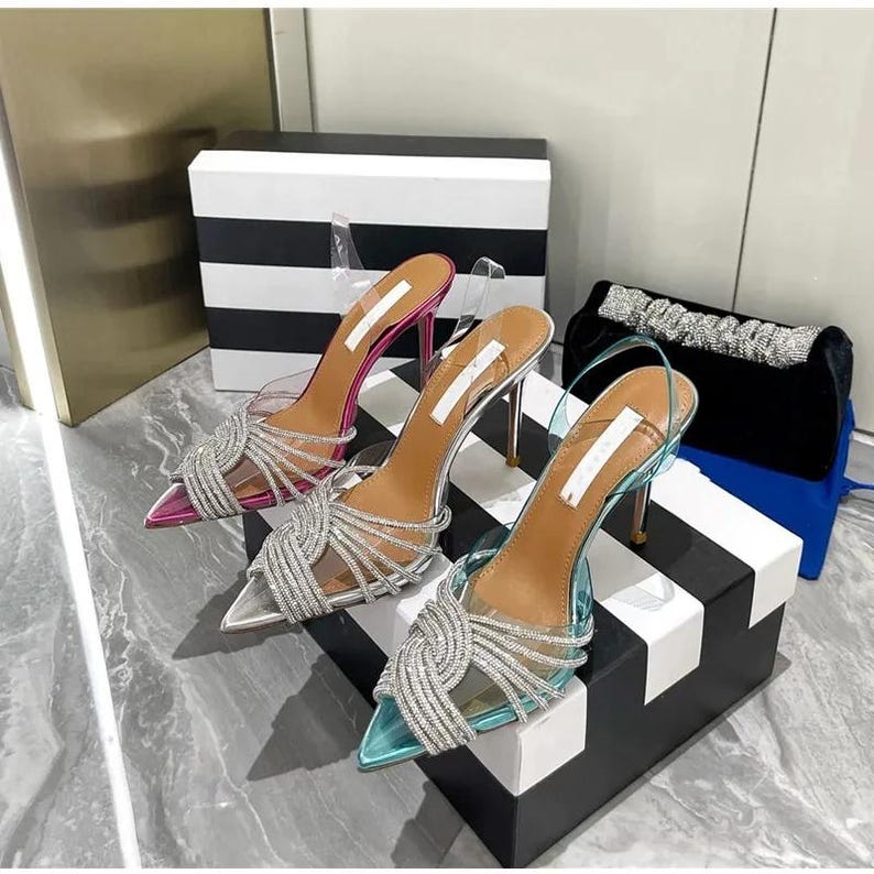 May include: Three pairs of high-heeled shoes in pink, silver, and teal. Each shoe has a pointed toe, clear straps, and a silver rhinestone design. The heels are thin and metallic. A black clutch with a silver beaded strap is also visible.