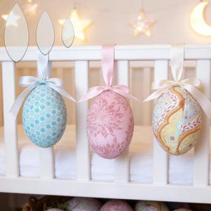May include: Three decorative Easter egg ornaments suspended from a white cot rail. The eggs are in pastel shades: blue with white spots, pink with a floral pattern, and a patterned egg with yellow and blue details. Each egg is tied with a satin ribbon.