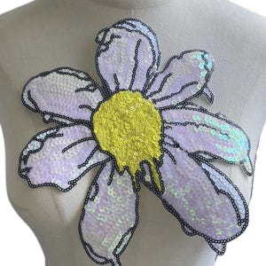 Könnte beinhalten: Eine große, paillettenbesetzte Blumenapplikation mit gelbem Zentrum und lavendelfarbenen Blütenblättern, die schwarz umrandet sind. Die Blume ist an einer Schaufensterpuppe befestigt und zeigt ihre Größe und ihr Design. Die Pailletten erzeugen einen schimmernden Effekt.