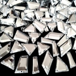 Puede incluir: Una colección de brillantes de cristal transparente facetado en diversas formas geométricas, incluyendo triángulos, rectángulos y trapecios. Los brillantes están dispersos sobre una superficie negra, reflejando la luz y creando un efecto brillante. Ideal para manualidades.