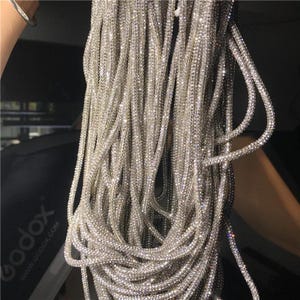 May include: Close-up of multiple strands of silver rhinestone trim. The trim is made of small, sparkling rhinestones strung together, creating a shimmering effect. The strands are hanging down, creating a cascading effect. The background is blurred.