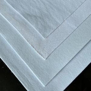 Pode incluir: Uma imagem em close-up de várias camadas de tecido branco, cada uma com uma textura ligeiramente diferente. As camadas são dispostas em um padrão diagonal, mostrando as variações na superfície do material, de lisa a sutilmente texturizada.