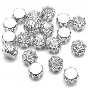 Può includere: Una collezione di abbellimenti in strass sfaccettati color argento. Gli strass sono rotondi con un aspetto chiaro e scintillante, incastonati in una base di metallo argentato. Gli abbellimenti sono sparsi su una superficie bianca.