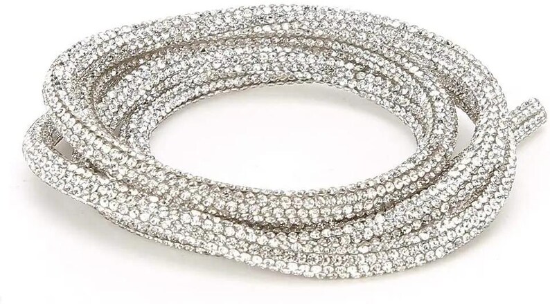 May include: A coiled, silver-colored necklace densely covered in small, sparkling rhinestones. The necklace is displayed against a white background, highlighting its reflective qualities and intricate design. The overall aesthetic is glamorous and eye-catching.