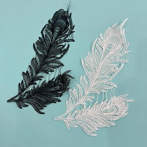 Puede incluir: Dos aplicaciones de encaje bordado con forma de plumas de pavo real, una en negro sólido y otra en blanco sólido. Cada pieza presenta un intrincado trabajo de hilo que detalla el ojo central y las delicadas barbas ramificadas del diseño.