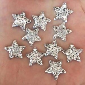 May include: Ten silver-coloured star-shaped embellishments, each covered in clear, faceted crystals. The crystals catch the light, creating a sparkling effect. These decorative stars are suitable for various craft projects and embellishments.