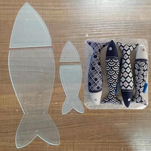 May include: A clear acrylic fish-shaped template with cut lines, next to four decorative fabric fish. The fabric fish have blue and white patterns, with white bellies and tail fins. The fish are arranged on a clear plastic base.