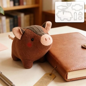 Peut inclure: Un cochon en peluche marron avec un museau, des oreilles et des joues rouges roses clairs. Un carnet marron et une étiquette sont à côté du cochon. Un patron du cochon est en arrière-plan.