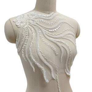 May include: A white lace appliqué featuring intricate embroidery, beads, and sequins arranged in a flowing, organic pattern. The design includes a floral cluster at the shoulder with trailing, curved lines of beadwork on a sheer mesh base.