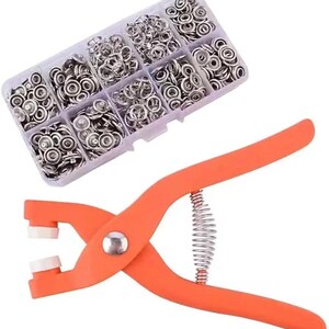 May include: An orange snap fastener tool with a silver spring and white tips, next to a clear plastic organizer filled with silver snap fasteners. The organizer is divided into compartments, each holding a different quantity of snaps.
