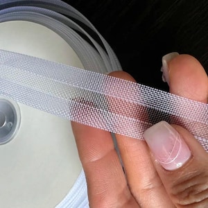 Op de afbeelding: Close-up van een rol wit elastisch lint. Het lint wordt in een hand gehouden en toont zijn geweven textuur en semi-transparante uiterlijk. Het lint wordt waarschijnlijk gebruikt voor naai- of knutselprojecten.