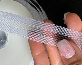 May include: Close-up of a roll of white elastic ribbon. The ribbon is held in a hand, showcasing its woven texture and semi-transparent appearance. The ribbon is likely used for sewing or crafting projects.
