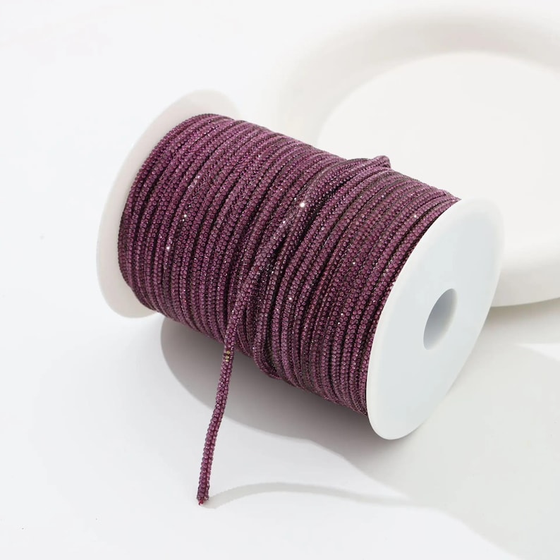 May include: A spool of dark purple, sparkly cord. The cord is tightly wound around a white plastic spool. The cord has a slight sheen, and the individual strands are visible. The cord is likely used for crafting or jewelry making.