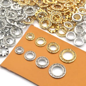 Könnte beinhalten: Eine Auswahl an Metallösen in Gold, Silber und klarem Kristall. Die Ösen sind rund mit einem zentralen Loch, einige mit einem sternförmigen Design. Sie sind auf einer braunen Oberfläche angeordnet, geeignet für Bastelarbeiten und Schmuckherstellung.