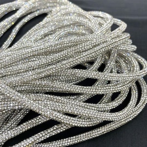 May include: Close-up of a bundle of silver rhinestone trim. The trim is made of small, sparkling rhinestones set closely together, creating a shimmering effect. The trim is flexible and appears to be on a black surface. Ideal for embellishing garments or accessories.