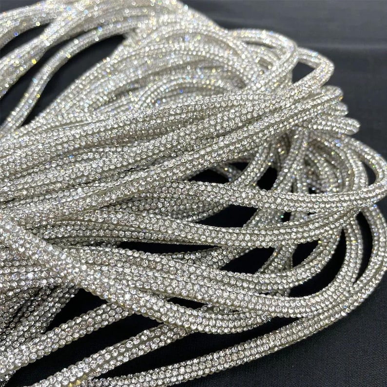 May include: Close-up of a bundle of silver rhinestone trim. The trim is made of small, sparkling rhinestones set closely together, creating a shimmering effect. The trim is flexible and appears to be on a black surface. Ideal for embellishing garments or accessories.