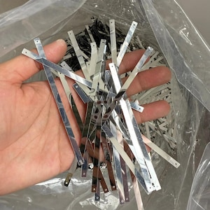 May include: A collection of thin, flat, silver-colored metal strips with a reflective finish. Each strip is rectangular and features a small, circular hole punched near both ends. The strips are loose and appear to be components for assembly.