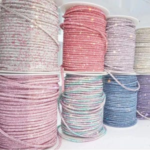May include: Spools of rhinestone trim in various colors, including pink, red, blue, and silver. The trim is tightly wound on white spools, creating a textured and sparkling appearance. Ideal for crafting and embellishments.