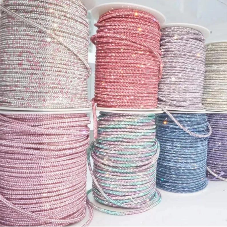 May include: Spools of rhinestone trim in various colors, including pink, red, blue, and silver. The trim is tightly wound on white spools, creating a textured and sparkling appearance. Ideal for crafting and embellishments.