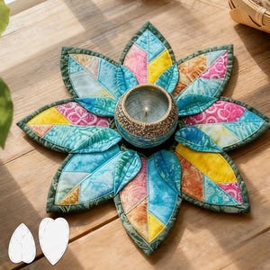 May include: A floral-shaped quilted candle holder. The petals are made from patchwork fabric in shades of blue, pink, yellow, and orange. A small, gold-toned candle holder with a lit candle is in the centre. It rests on a wooden surface.