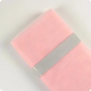 Può includere: Un pezzo piegato di tessuto in tulle rosa chiaro caratterizzato da una trama a rete fine e semi-trasparente. Il tessuto è fissato al centro da una fascia di carta grigio chiaro, liscia e solida, che avvolge orizzontalmente il materiale.