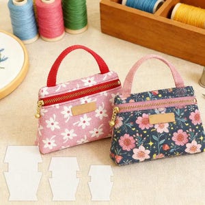May include: Two small fabric pouches with top handles and metal zippers. One pouch is pink with a white floral pattern and a red handle, while the other is dark blue with a pink floral pattern and a pink handle. Both feature a blank rectangular label.