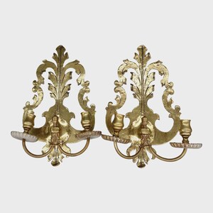 May include: A pair of gold-toned metal wall sconces featuring ornate, leaf-like scrollwork backplates. Each sconce has two curved arms extending outward, supporting candle cups with scalloped, textured drip pans at the base of each holder.