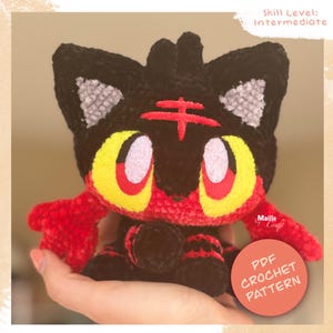 Peut inclure: Une peluche au crochet représentant un personnage félin noir et rouge avec de grands yeux jaunes, des oreilles intérieures gris clair et une marque rouge sur le front. Le texte "Skill Level: Intermediate" et "PDF CROCHET PATTERN" est visible.
