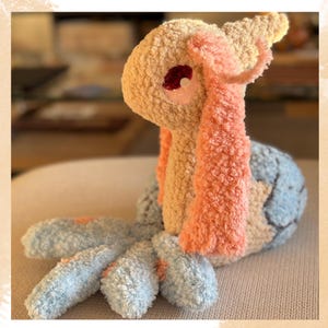 Puede incluir: Un peluche artesanal con forma de criatura, con cabeza beige, ojo rojo y detalles rosas. El cuerpo es azul y beige, con ap&eacute;ndices azules y rosas. Fabricado con tejido suave y texturizado.