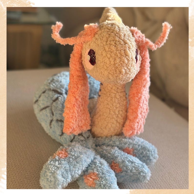 Puede incluir: Un peluche hecho a mano con forma de criatura marina. El juguete tiene un cuerpo beige claro, orejas y detalles rosas y tent&aacute;culos azules. Los ojos son de color rojo oscuro. El peluche est&aacute; hecho de tela suave y texturizada.