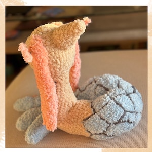 Puede incluir: Un peluche hecho a mano con forma de criatura, con un cuerpo melocot&oacute;n claro, brazos rosas y una cola estampada en azul y gris. El juguete est&aacute; hecho de hilo suave y texturizado, lo que le da un aspecto tierno.