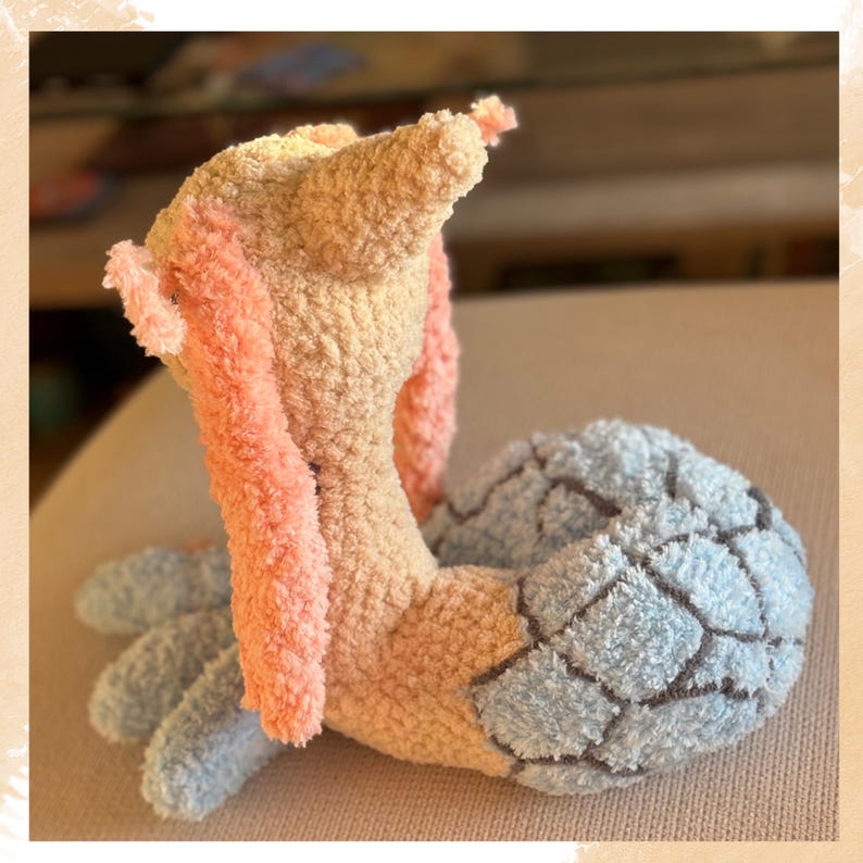 Puede incluir: Un peluche hecho a mano con forma de criatura, con un cuerpo melocot&oacute;n claro, brazos rosas y una cola estampada en azul y gris. El juguete est&aacute; hecho de hilo suave y texturizado, lo que le da un aspecto tierno.