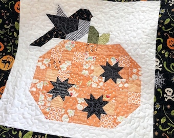 May include: A white quilted fabric square with a black bird and an orange pumpkin made of patchwork fabric. The pumpkin has black stars and a green stem. The fabric has a floral pattern.