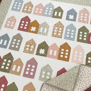 May include: A white quilt with a patchwork design featuring various coloured houses. The houses are arranged in rows and have different patterns and colours, including red, blue, green, and yellow. The quilt is made with a variety of fabrics and textures.