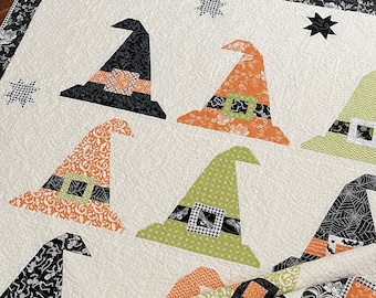 May include: A white quilt with a Halloween theme featuring various witch hats in different colors and patterns. The hats are made of fabric and have a variety of embellishments, including buckles, bows, and stars.