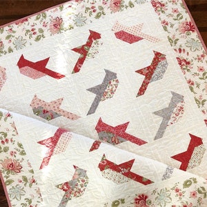 May include: A white quilt with a floral border and a patchwork design featuring red, pink, and gray birds. The quilt is made with a variety of fabrics and has a quilted texture.