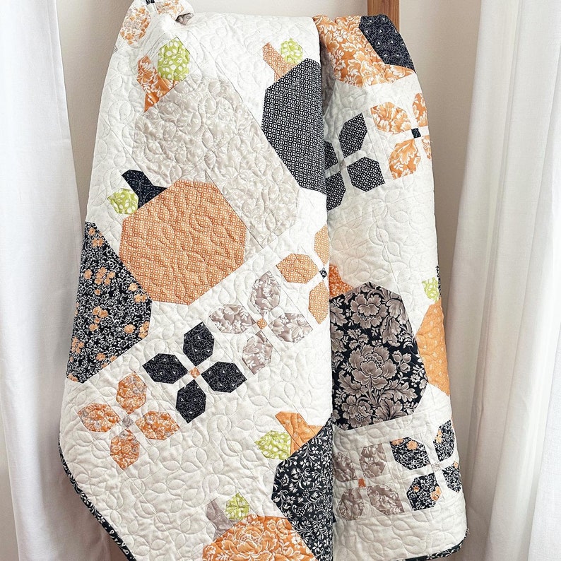 May include: A white quilt with a patchwork design featuring black, orange, and green fabric. The quilt is made up of various geometric shapes and patterns.