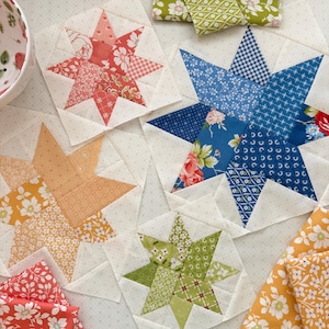 May include: Four fabric quilt blocks featuring star patterns in coral, blue, yellow, and green floral and geometric prints. Each block is constructed from pieced cotton fabric squares on a light cream background with small grey dots.